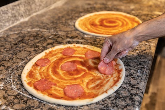 A hand placing pepperoni slices on pizza dough with tomato sauce.