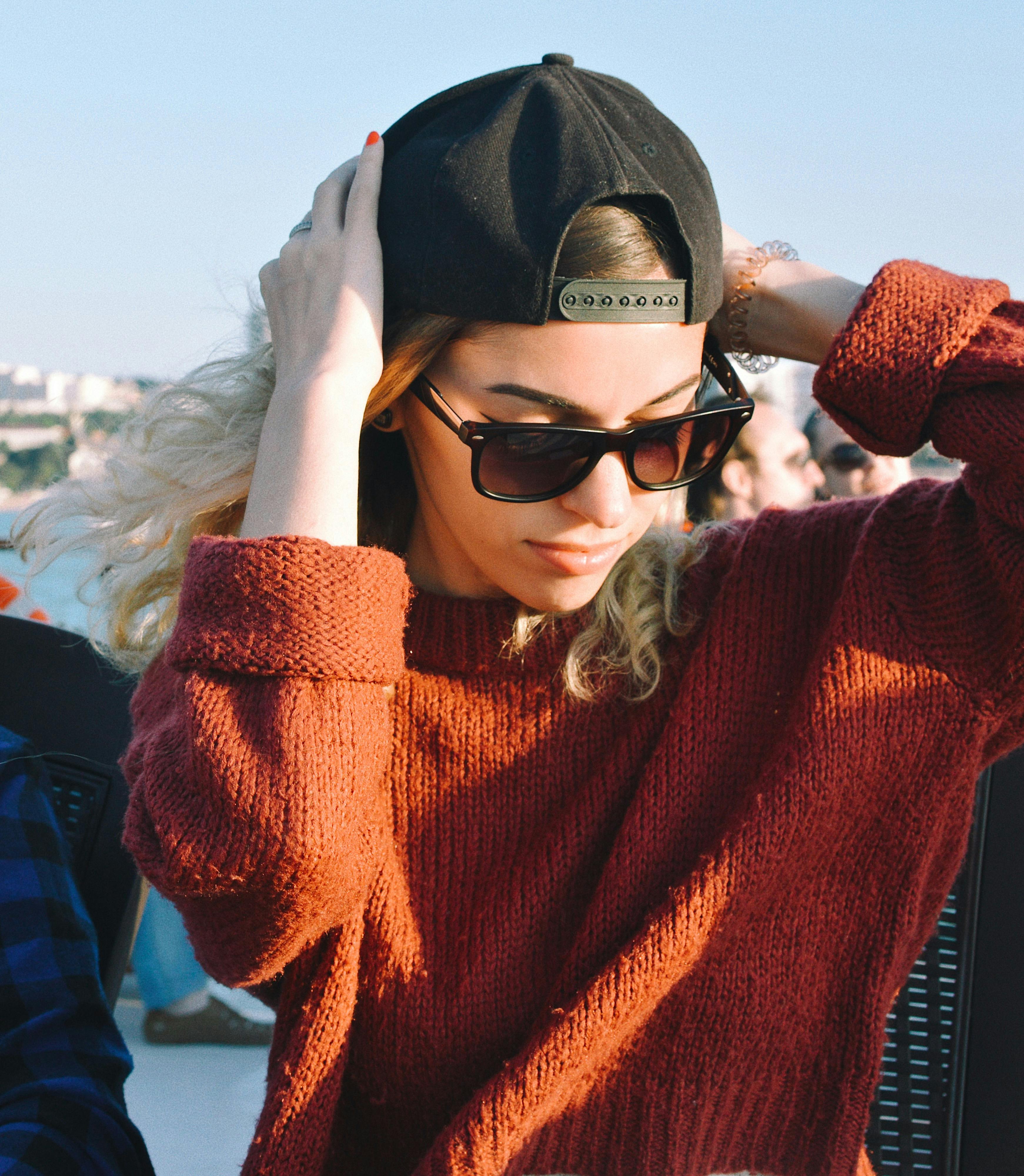 Fashionable young woman in a knit sweater adjusts her cap, enjoying sunny weather outdoors.