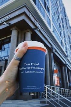 Hand holding a blue coffee cup with a motivational message in front of a building.