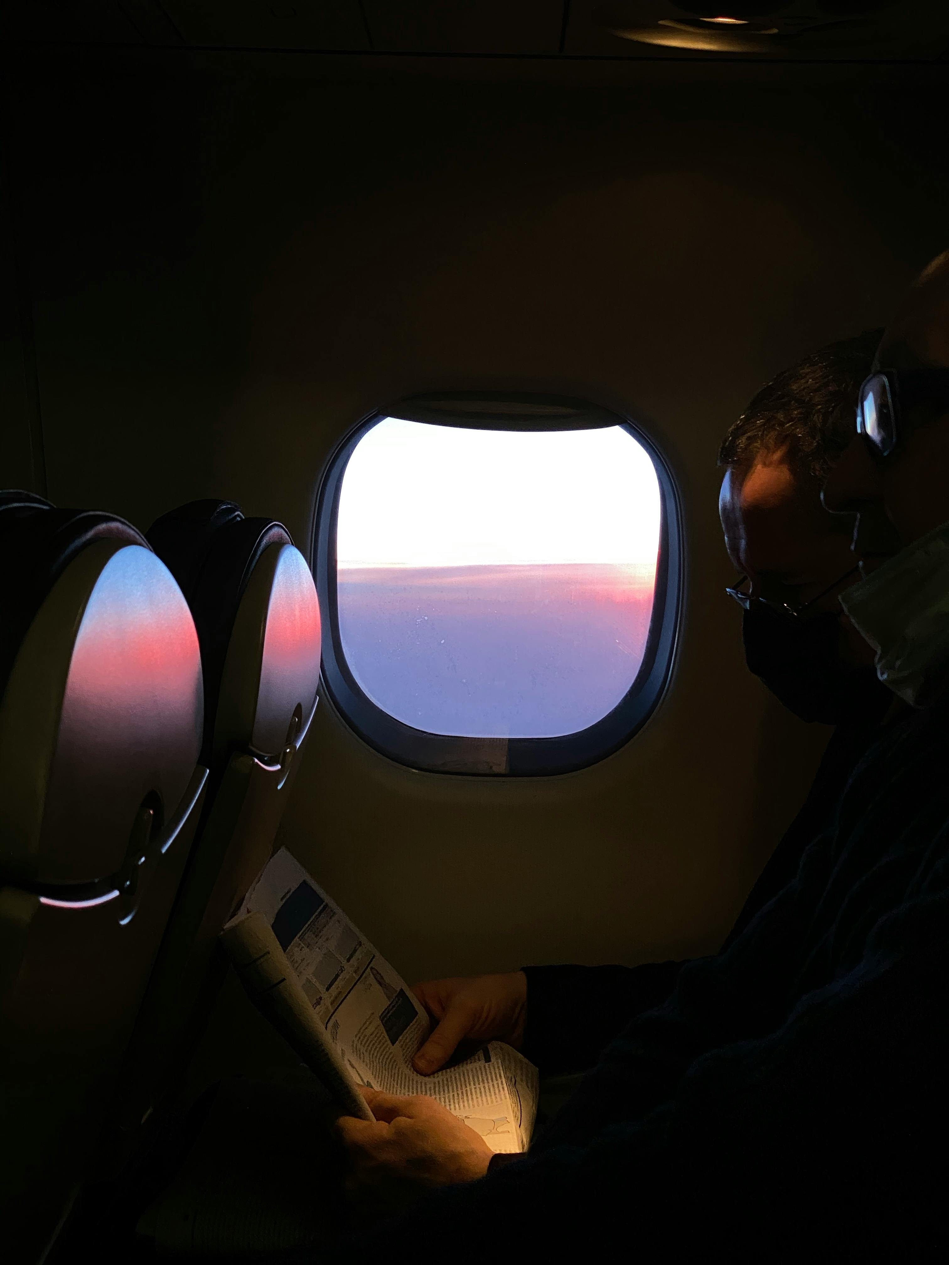 A passenger reads a magazine beside a sunset view from the airplane window, capturing a serene travel moment.