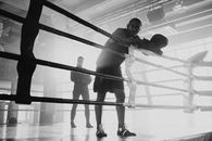 Grayscale Photo of a Man in Sportswear Holding Boxing Pads Leaning on Boxing Ring Ropes