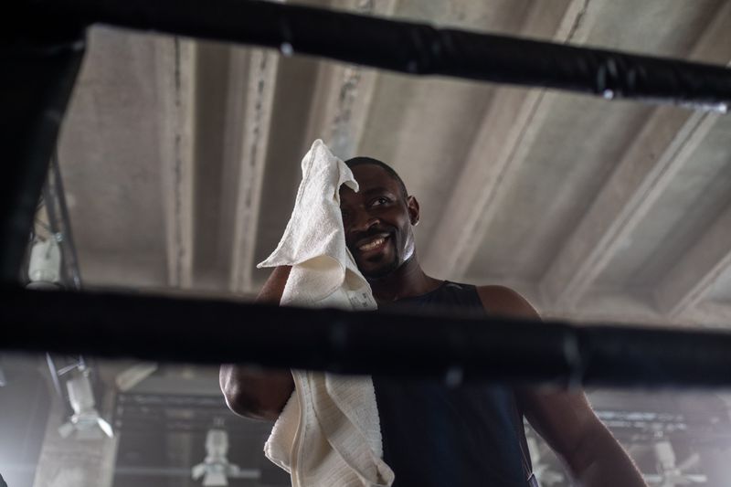 A Black man in a dark sleeveless top stands in a gym boxing ring, smiling as he wipes sweat from his face with a white towel. Sweat, heat, and friction are what inflame hair follicles after a workout.