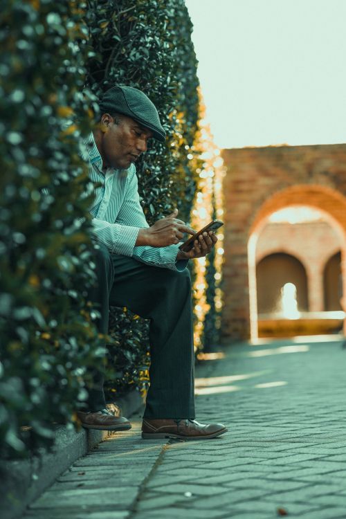Free A senior man with a cap is sitting outdoors, engaged with his smartphone on a sunny day. Stock Photo