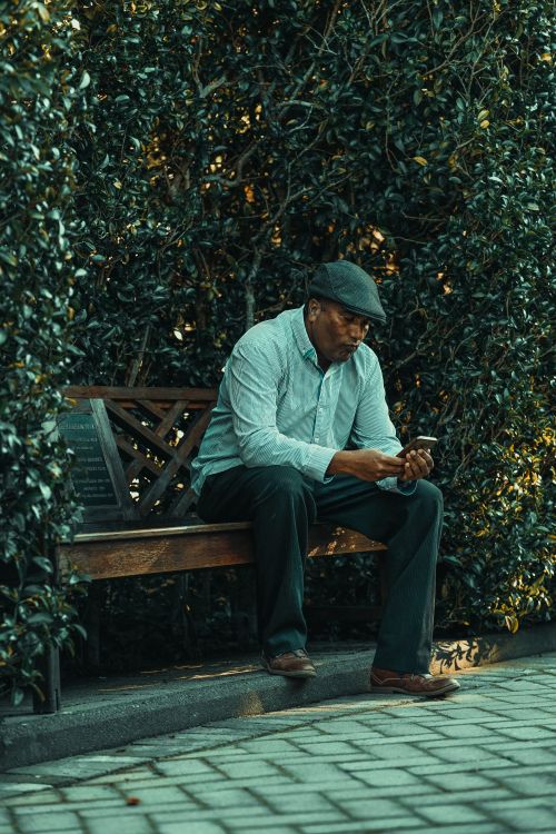 Free Senior African American man sitting on a park bench using his mobile phone in an outdoor garden setting. Stock Photo