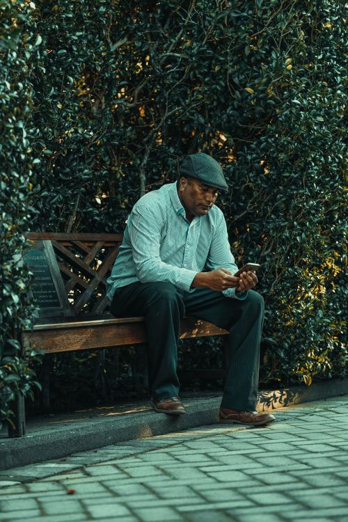 Free An older man using his smartphone while sitting on a wooden bench outdoors. Stock Photo