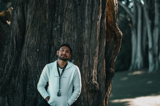 Casual man leaning against a large tree trunk in a peaceful outdoor setting.