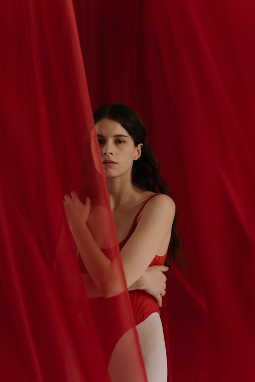 Young Woman In Red Swimsuit Standing Behind A Red Sheer Curtain Fabric