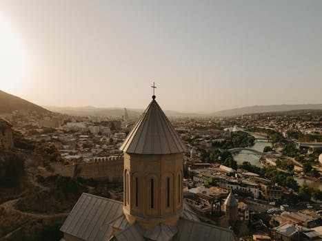 Aerial view of a church tower at sunset with the sprawling cityscape of Tbilisi, Georgia in the background.