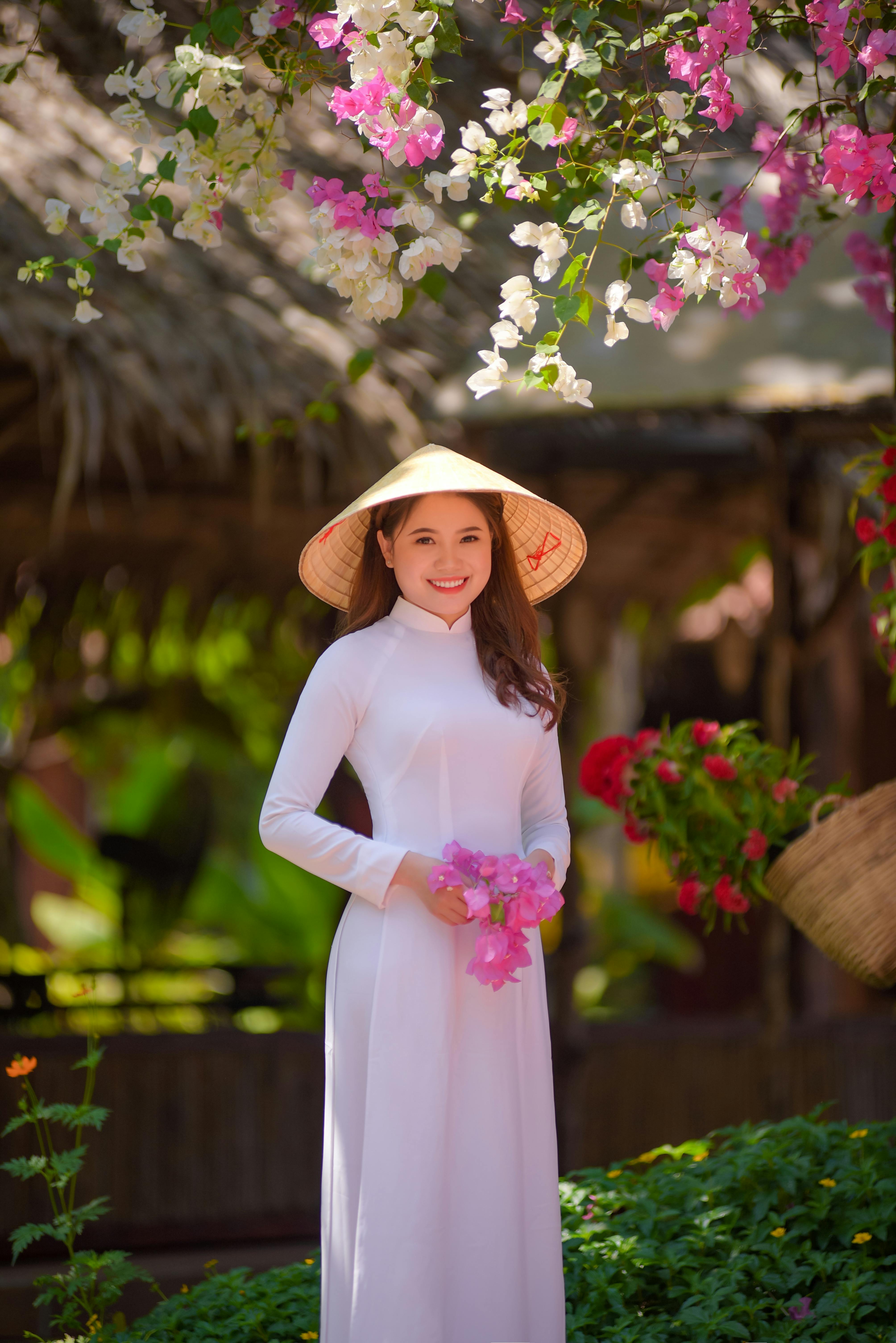 Smiling woman in traditional Ao Dai and conical hat holding flowers in a vibrant Vietnamese garden.
