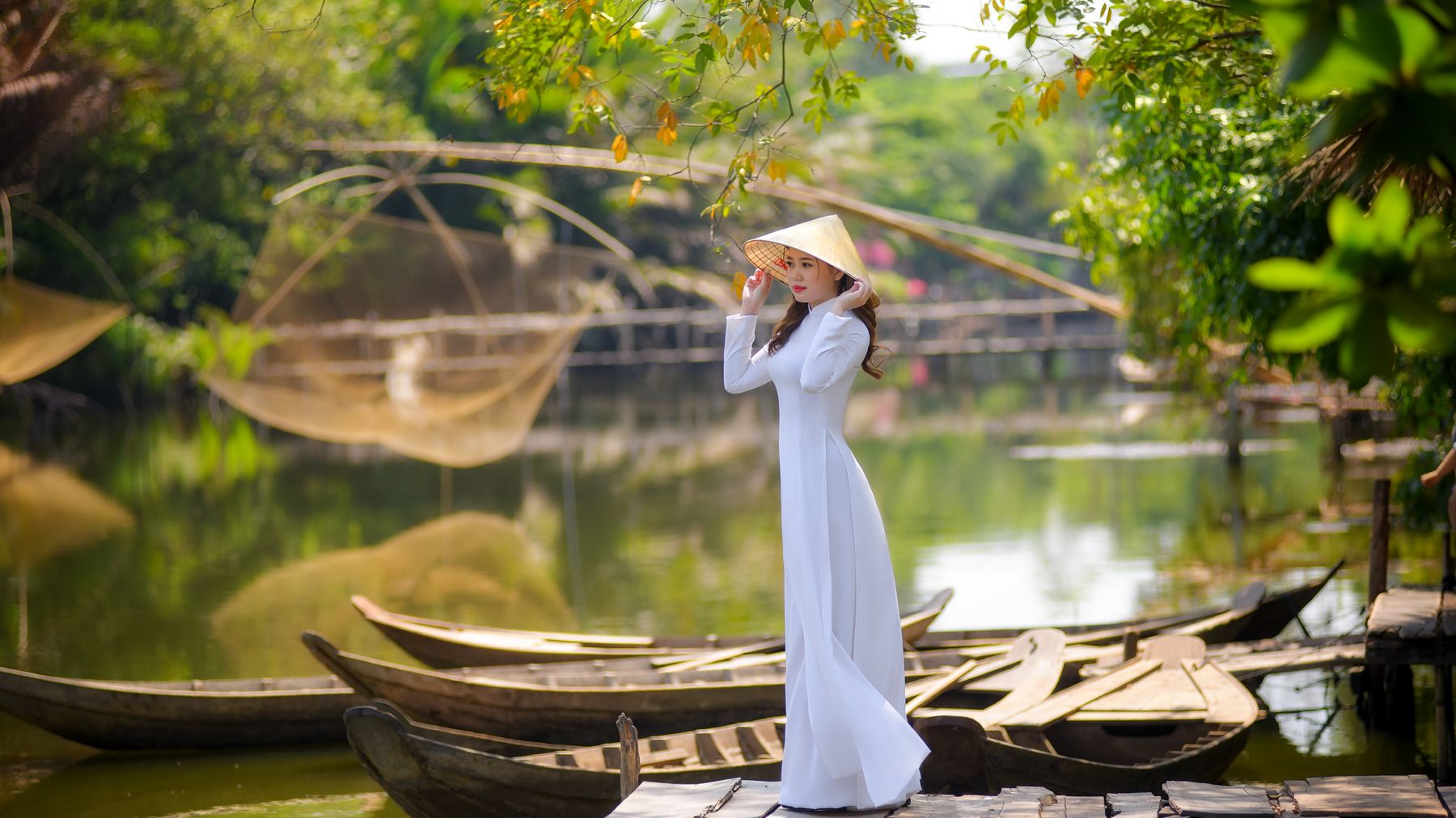 Elegant Vietnamese woman in ao dai with a non la by a serene pond, surrounded by lush greenery and traditional boats.
