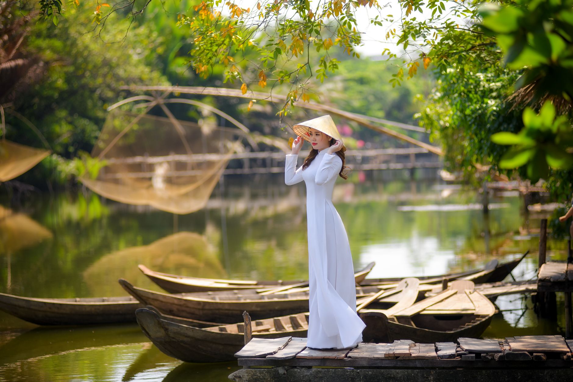 Elegant Vietnamese woman in ao dai with a non la by a serene pond, surrounded by lush greenery and traditional boats.