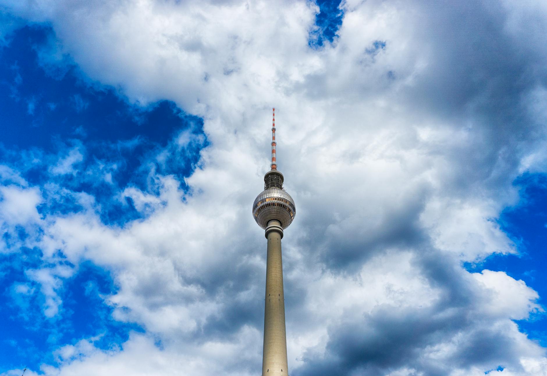 A dramatic view of the Berlin TV Tower (Fernsehturm) against a vibrant blue sky with scattered clouds.