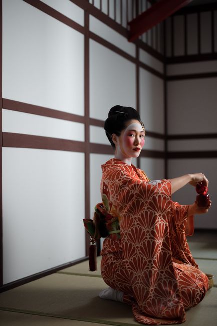 A Japanese geisha kneeling in a room, holding an item, showcasing traditional attire and culture.