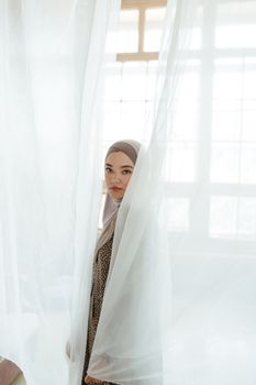 Elegant woman in hijab poses behind sheer white curtains with soft light filtering through.