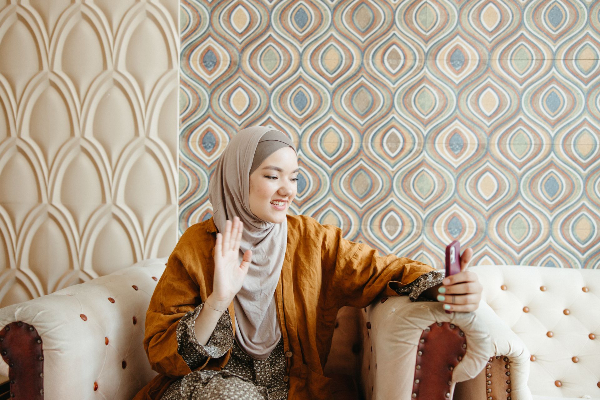Middle Eastern woman in a hijab smiling and waving during a video call indoors.