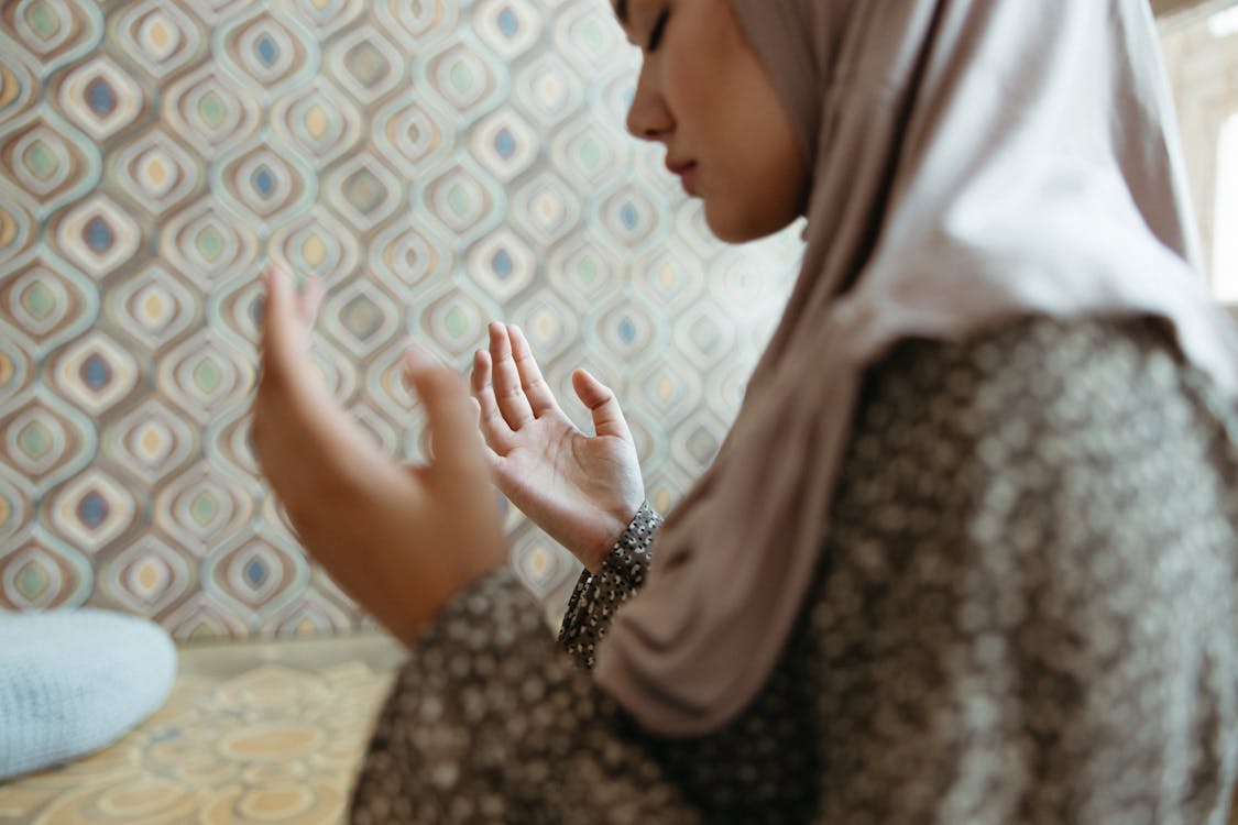 A young Muslim woman in a hijab meditates in a peaceful, patterned indoor setting.