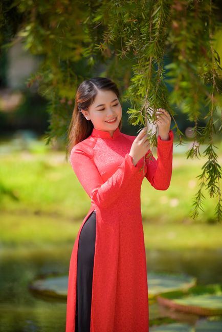 Smiling woman in red ao dai gently touching leaves in a serene garden setting.
