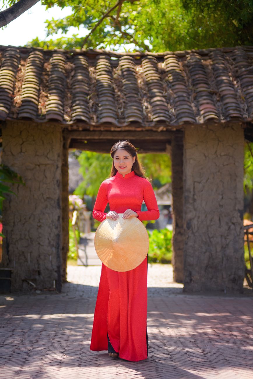 Asian woman in red Ao Dai holding conical hat, smiling in a sunny Vietnamese village setting.