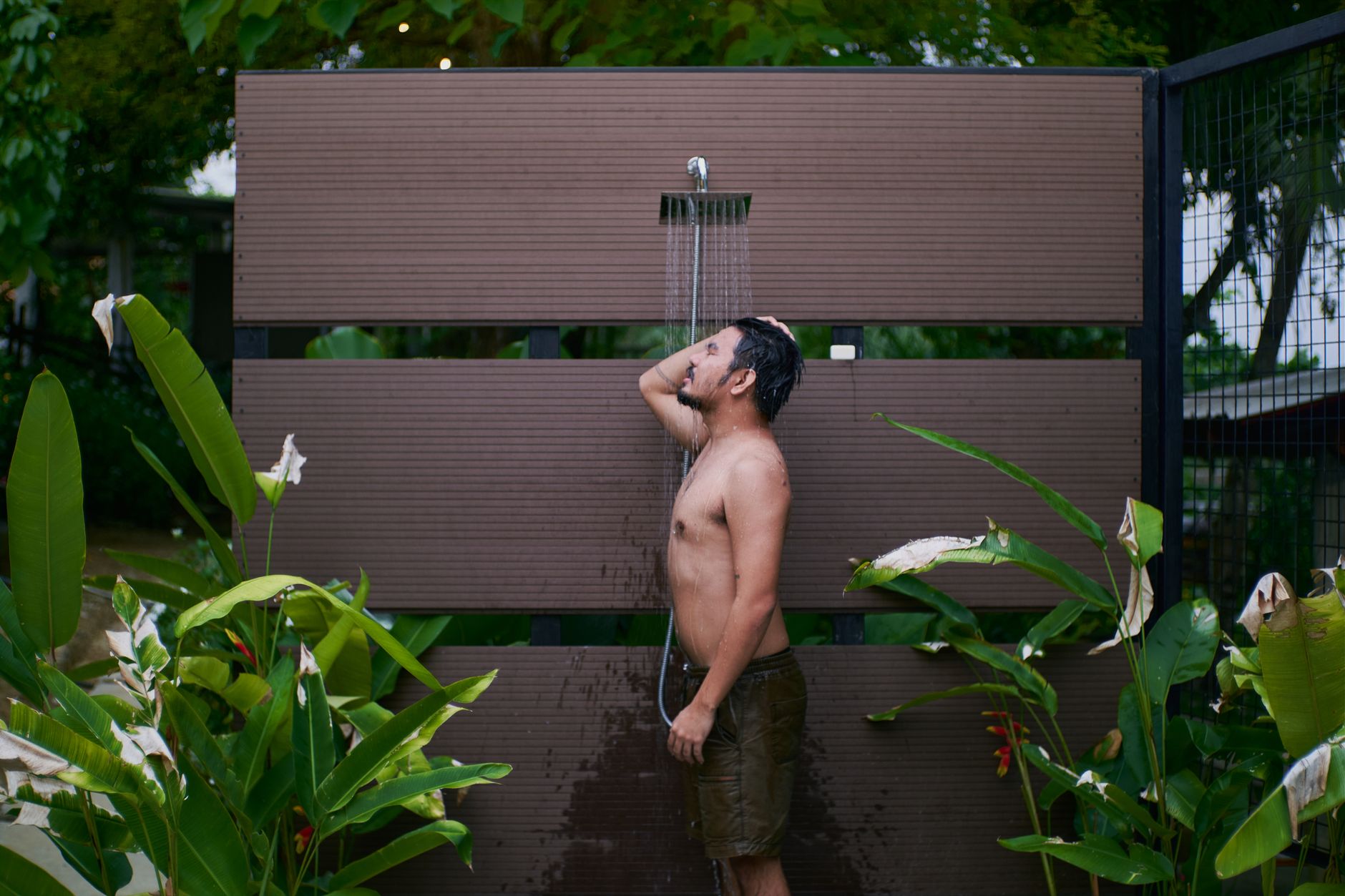 Young man enjoying an outdoor shower surrounded by lush tropical plants.