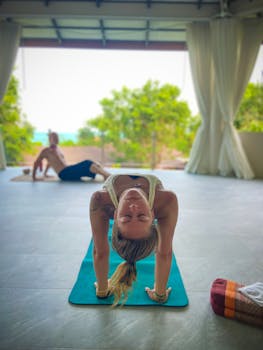 A woman performs a bridge yoga pose on a mat in an outdoor class setting with trees in the background.