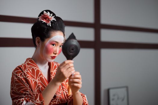 Geisha in traditional attire applying makeup with a hand mirror, reflecting Japanese culture.