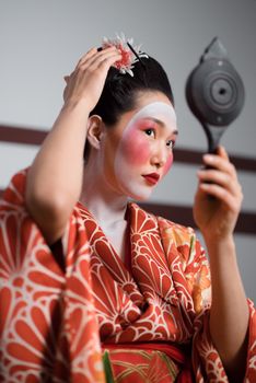 A Japanese geisha in traditional attire adjusts her hair while holding a hand mirror.