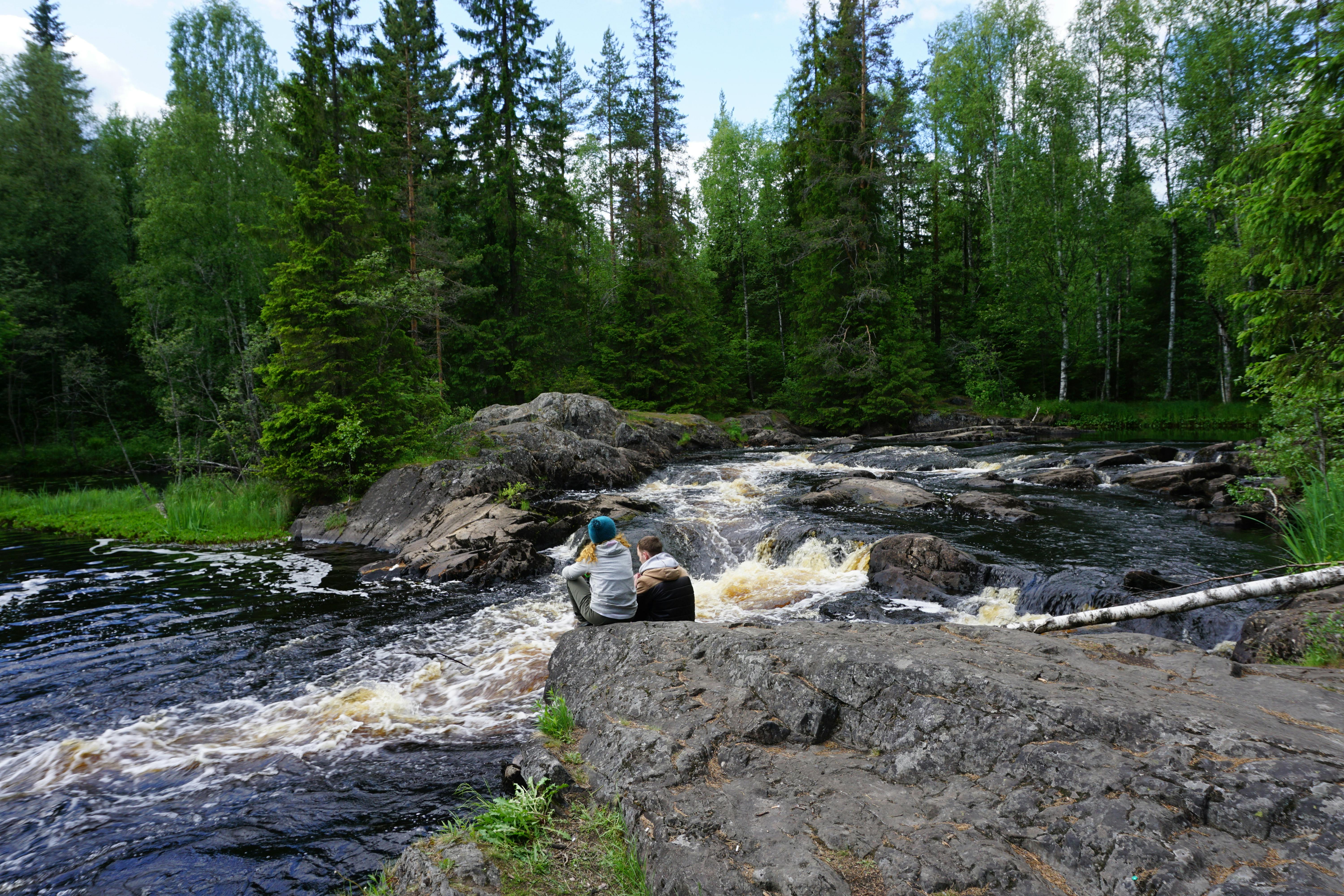 A couple sits on rocks by a flowing river in a lush, green forest, soaking in nature's serenity.