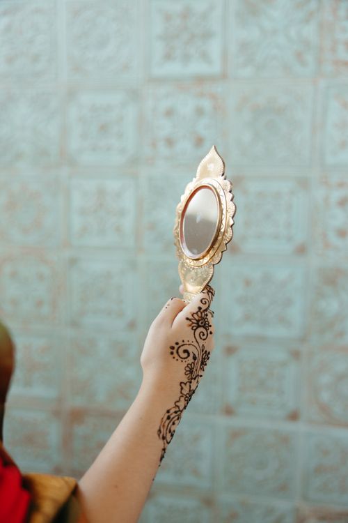 Free A hand with intricate henna designs holds an ornate mirror against a patterned background. Stock Photo