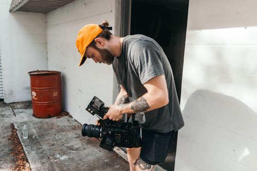 Free A tattooed videographer wearing an orange cap adjusts his camera equipment outdoors, preparing for a video shoot. Stock Photo