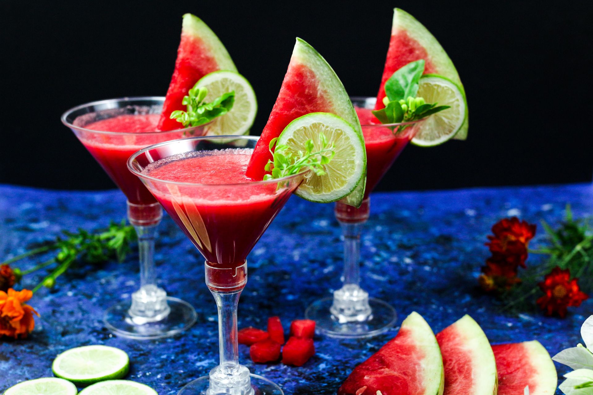 Colorful watermelon and lime mocktails on a blue background with fresh fruit garnishes.