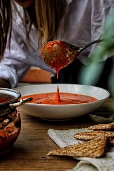A ladle pours fresh tomato soup into a bowl, surrounded by crisp bread.