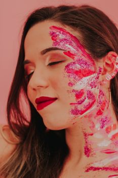 Close-up of a woman with artistic face paint in a vibrant, creative style on a pink background.