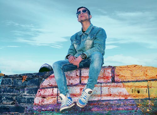 Man in Gray Denim Jacket and Jeans Sitting on Brick Wall