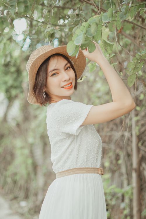 Free Asian woman smiling in a white dress and sun hat, standing outdoors in lush greenery. Stock Photo
