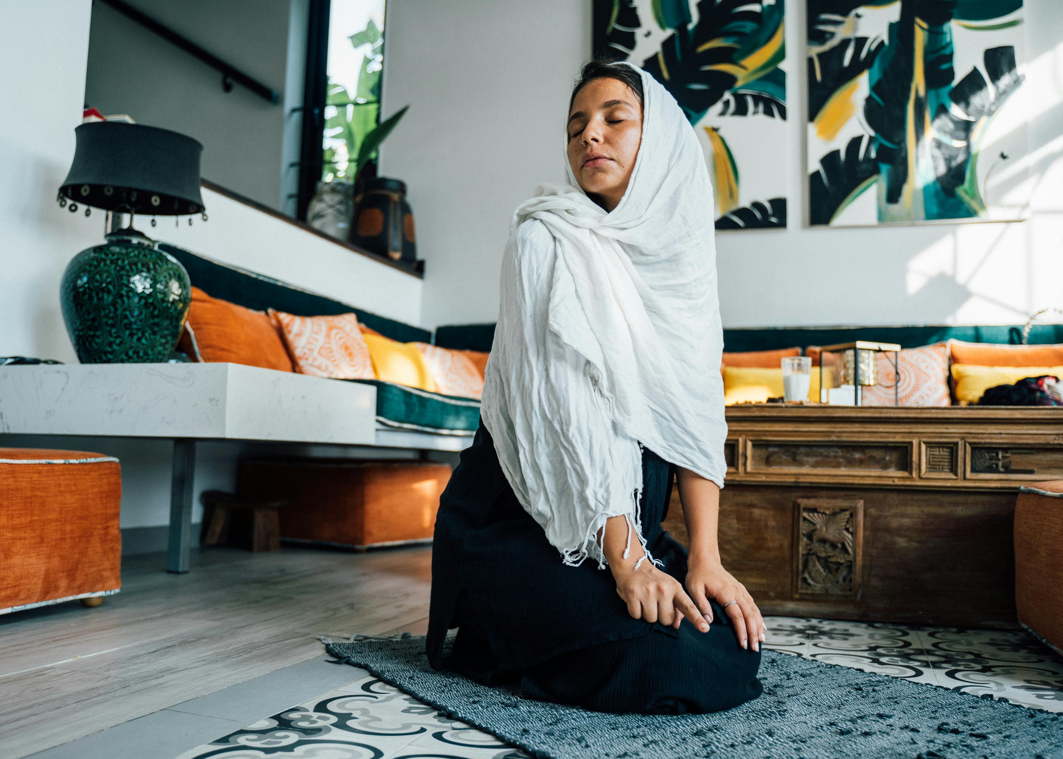 A Muslim woman in a hijab kneeling and praying on a decorative rug indoors.