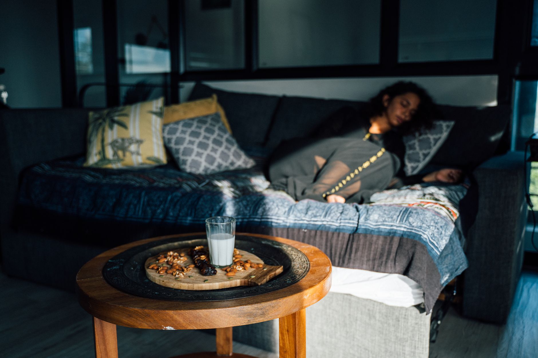 A woman naps on a sofa in a sunlit room with snacks and a glass of milk on a table.