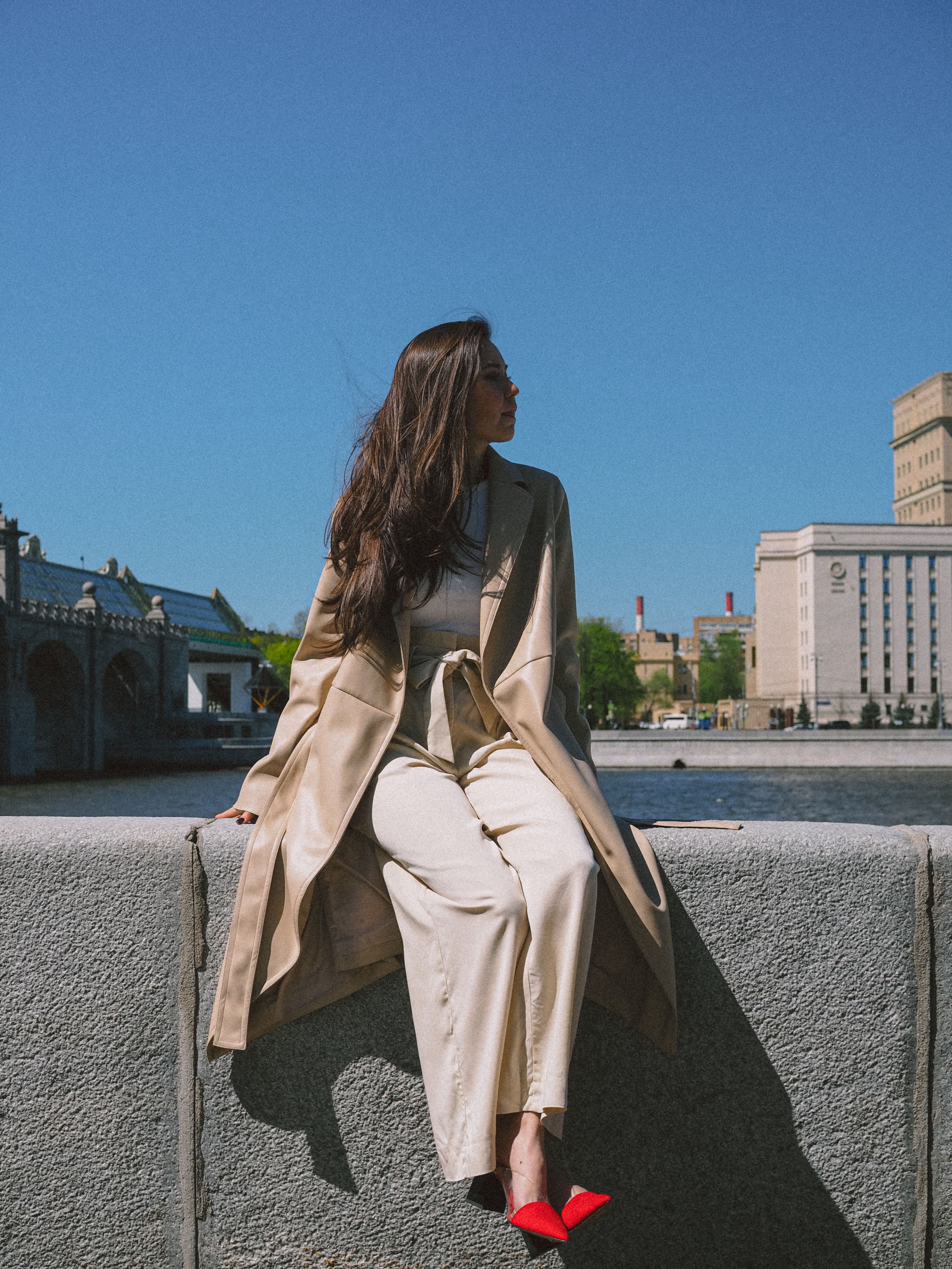 Stylish woman in beige coat sits by riverside in urban cityscape.