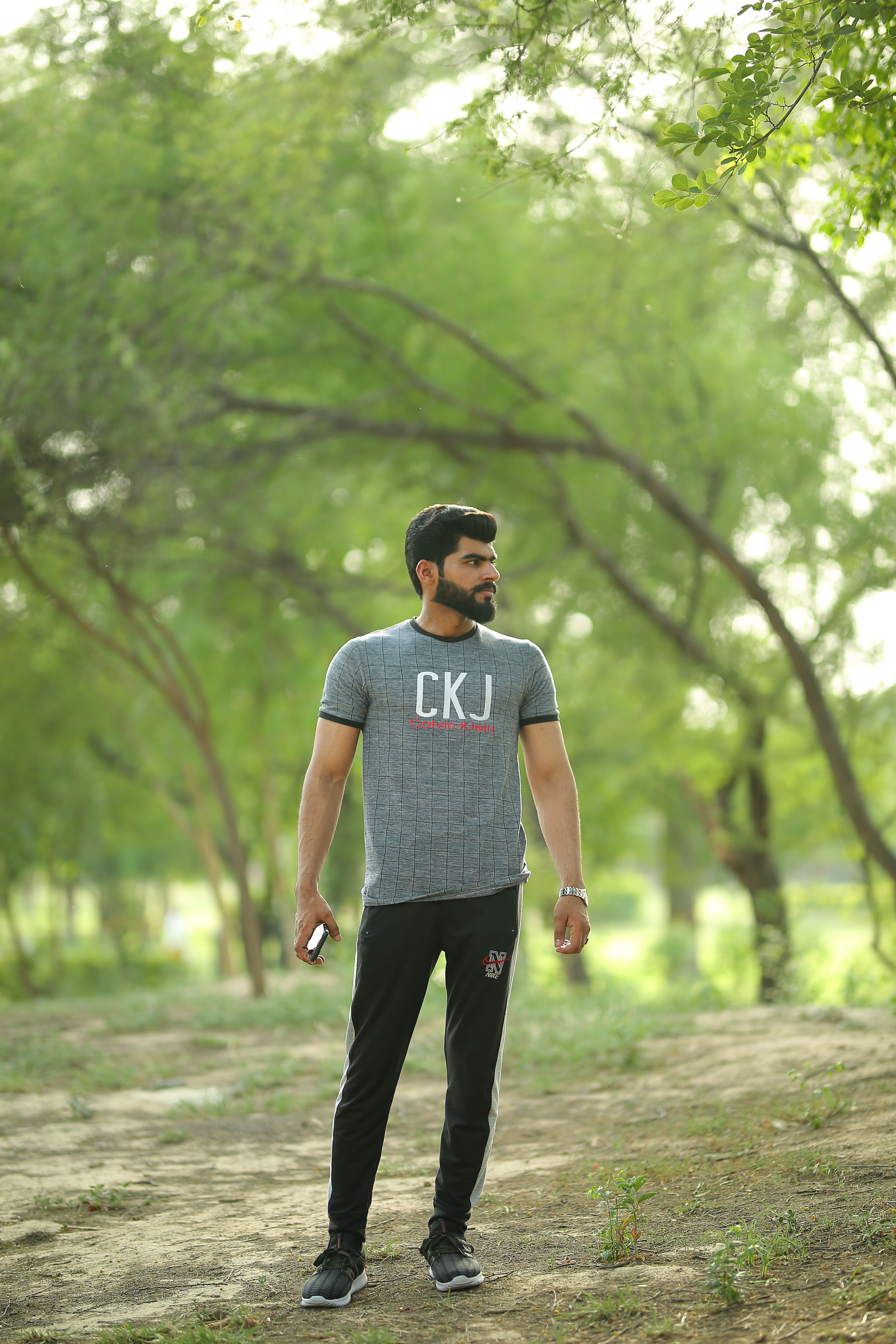 A bearded man stands outdoors in a lush park, wearing a gray t-shirt with 'CKJ' print.