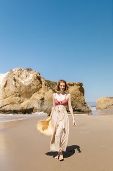 A smiling woman walks barefoot on the sandy beach holding a straw hat on a sunny day.