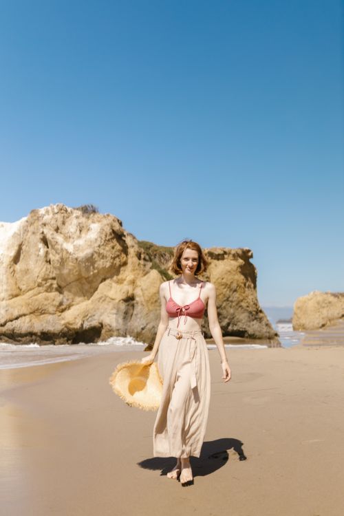 Free A smiling woman walks barefoot on the sandy beach holding a straw hat on a sunny day. Stock Photo