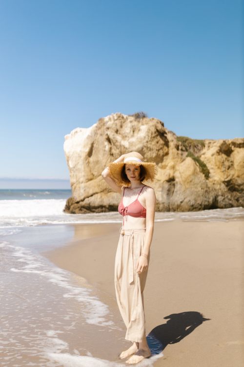 Free A young woman stands on a sandy beach during a sunny day, wearing a straw hat. Stock Photo