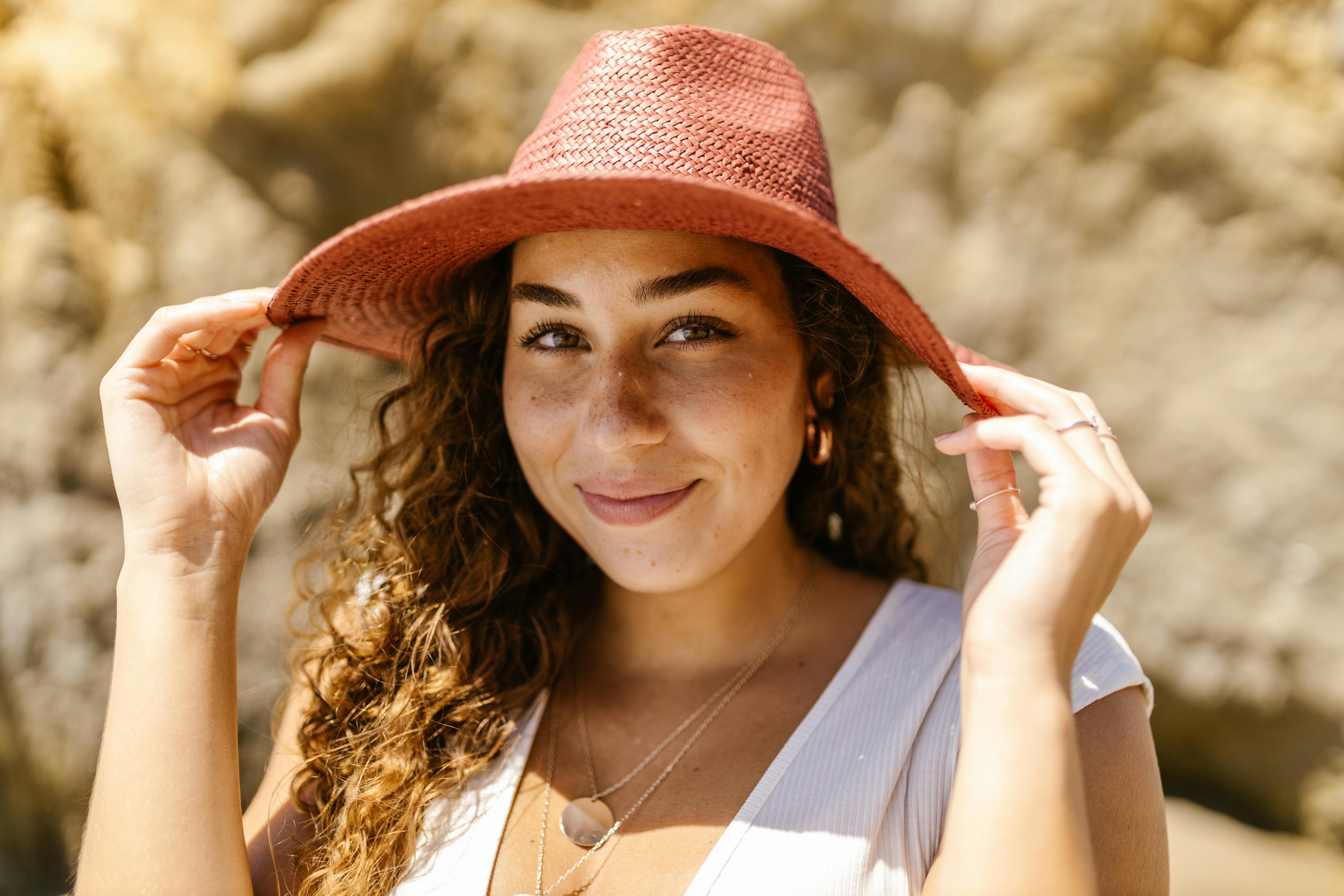 A young woman with curly hair smiling while holding a sun hat outdoors.