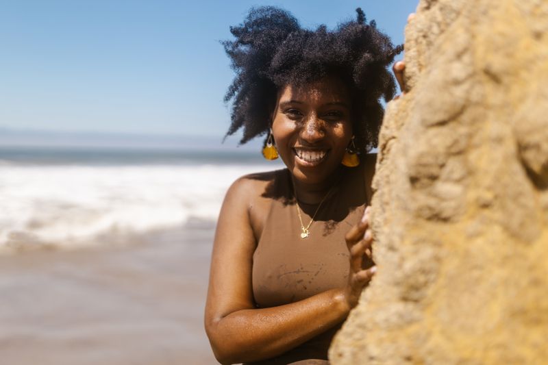 A Black woman with natural hair smiles in full sun at the beach, bare shoulders in direct light.