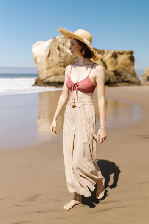 Free A woman in a straw hat and bikini top walks along a sunny beach shore. Stock Photo