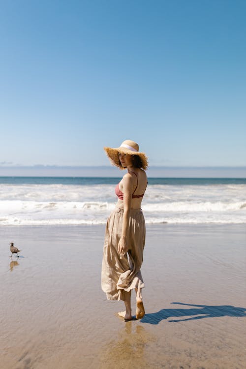 A Woman with Straw Hat Walking on Wet Shore