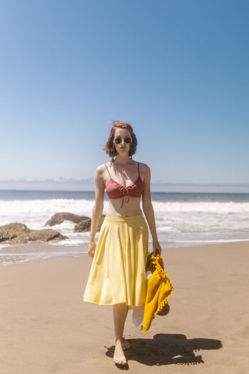 Free Woman in a bikini top and skirt walking on a sunny beach with the sea in the background. Stock Photo