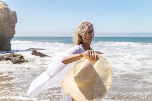 Smiling Woman Walking on the Beach Holding a Straw Hat 