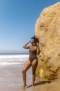 Confident woman in swimsuit posing near a rock on a sunny beach, enjoying summer vacation.