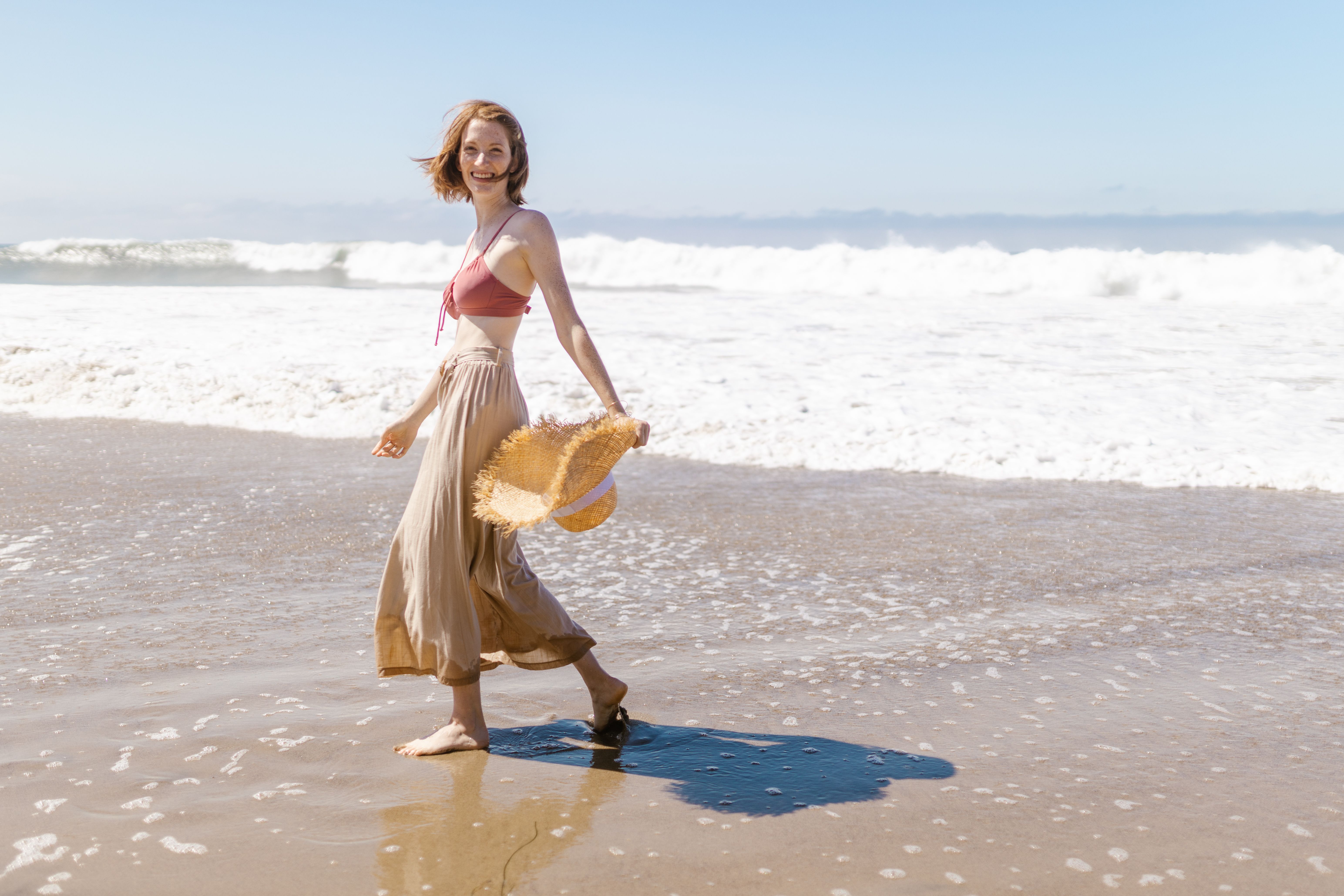Smiling Woman Walking on the Beach Holding a Straw Hat · Free Stock Photo