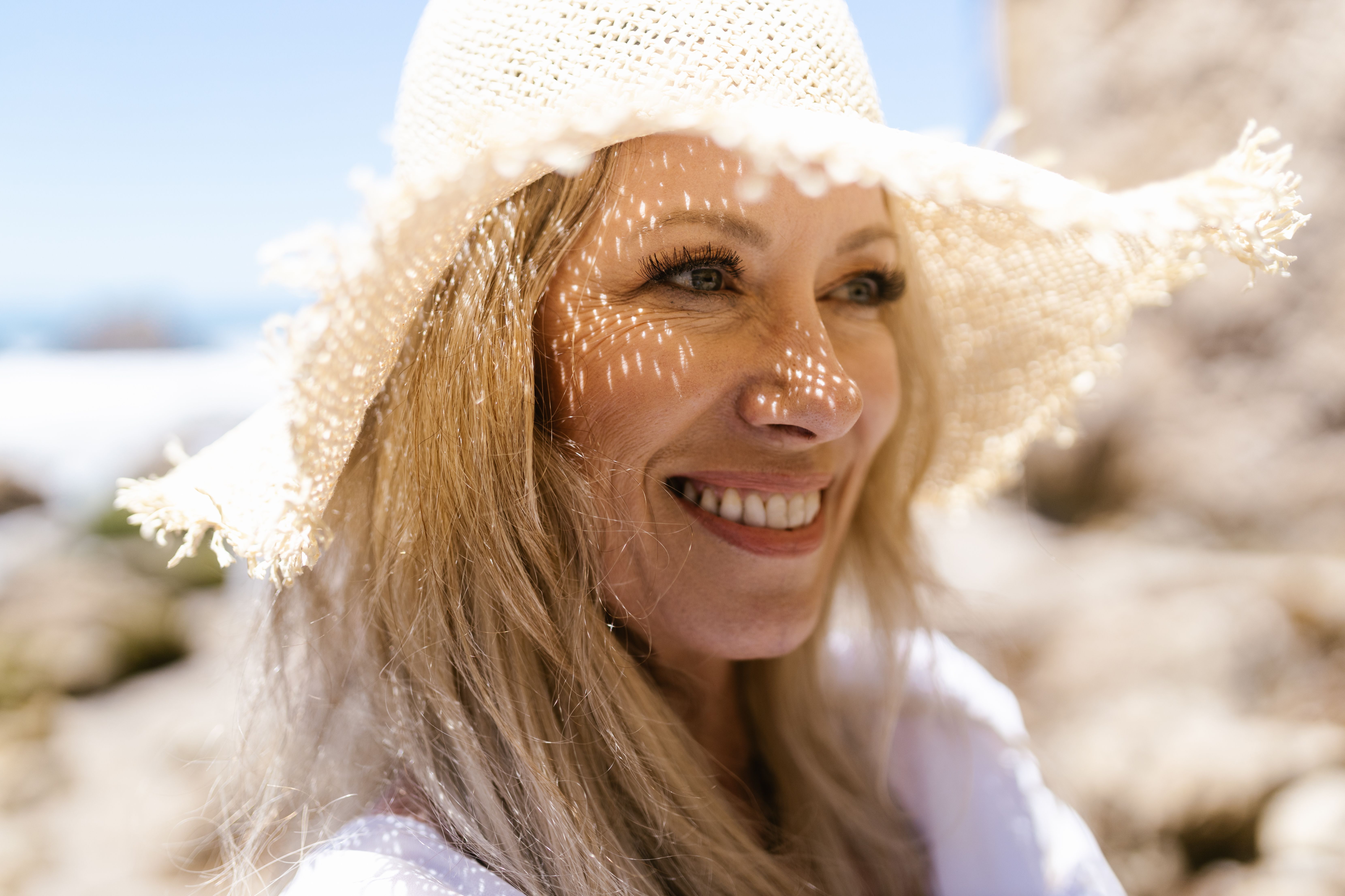 Close-up of a smiling woman wearing a sun hat at the beach. Bright and cheerful summer vibe.